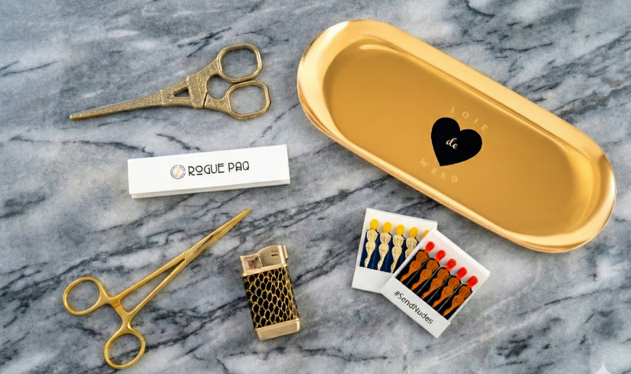 A luxury cannabis gift set featuring a booklet of Rogue Paq unbleached hemp rolling papers, Eiffel Tower Scissors, a refillable lighter, two books of SendNudes matches, and a pair of gold-toned hemostat jay clips. The set is presented in a minimalist, high-contrast arrangement that highlights the polished gold hardware.