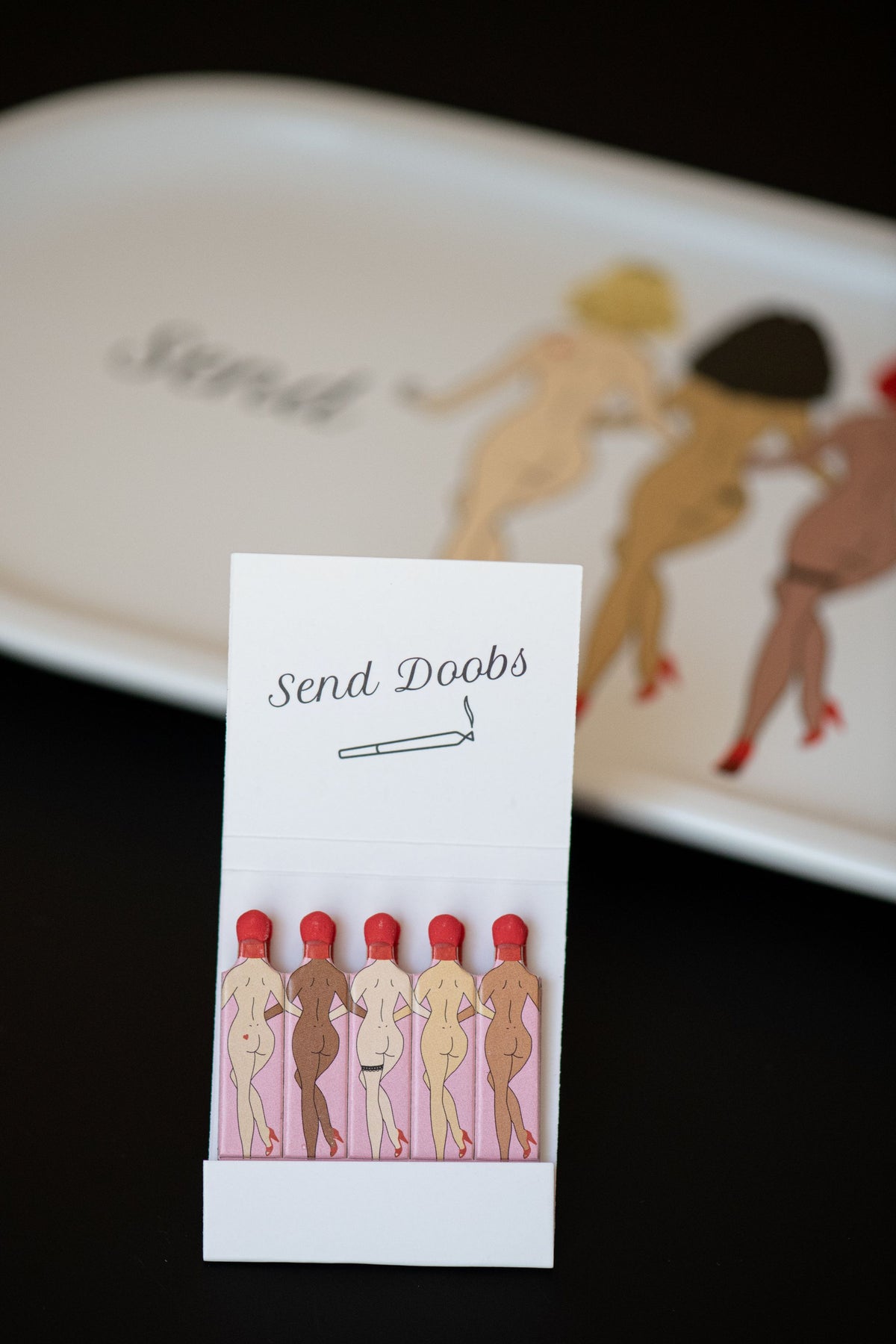 Single book of Rogue Paq Send Doobs Matches highlighting the detailed 'Send Doobs' illustration with vibrant colors and retro typography set in front of the Send Doobs Valet Rolling Tray.