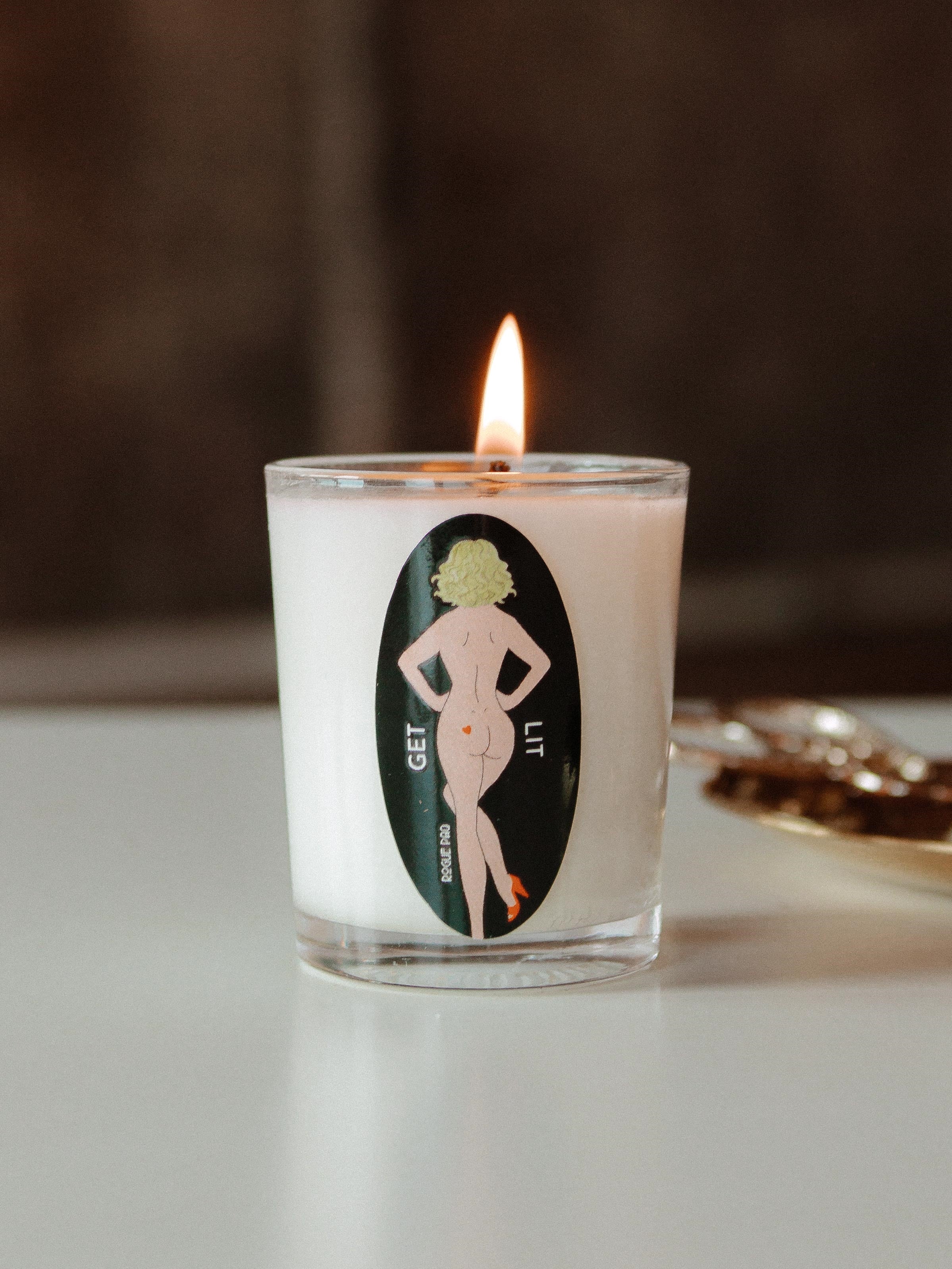 A minimalist clear glass votive candle with cheeky sticker filled with off-white organic soy wax. The label features the signature SendNudes dancing nude and the Rogue Paq logo and the playful 'GET LIT' and 'SendNudes' typography in a sophisticated black font. A single hemp wick is centered in the hand-poured wax. Part of the lineup of cute smoking accessories.