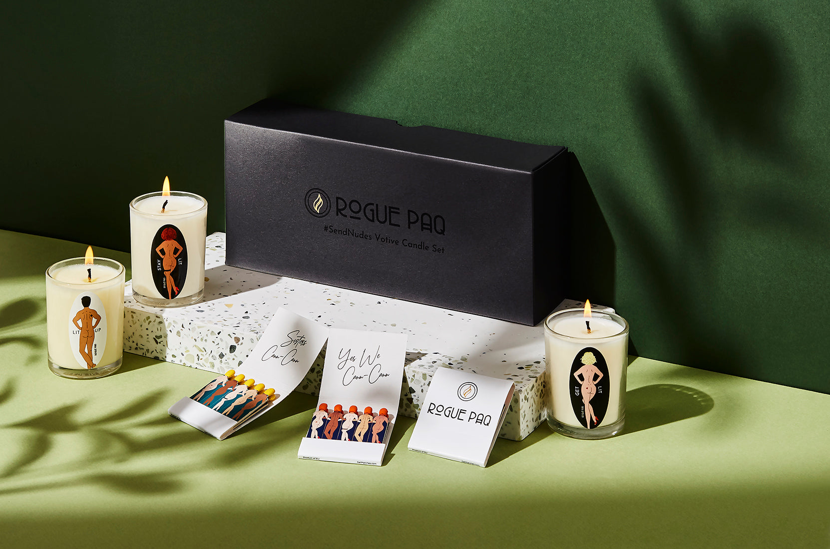A detailed shot of three soy wax aromatherapy candles plue Rogue Paq designer matchbooks from the SendNudes collection. The 'Sisters Cann-Cann'  and Yes We Cann Cann illustrations feature diverse, artistic nude figures with varying skin tones and hair colors in a high-kick dance pose, representing a celebration of body positivity and inclusivity. The shot includes the sleek minimalist matte black gift box.